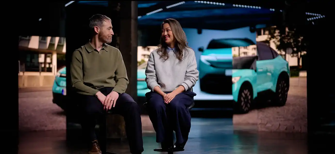 Chris and Julie Ramsey sit on stools in front of a turquoise car, smiling at each other in a dark studio.