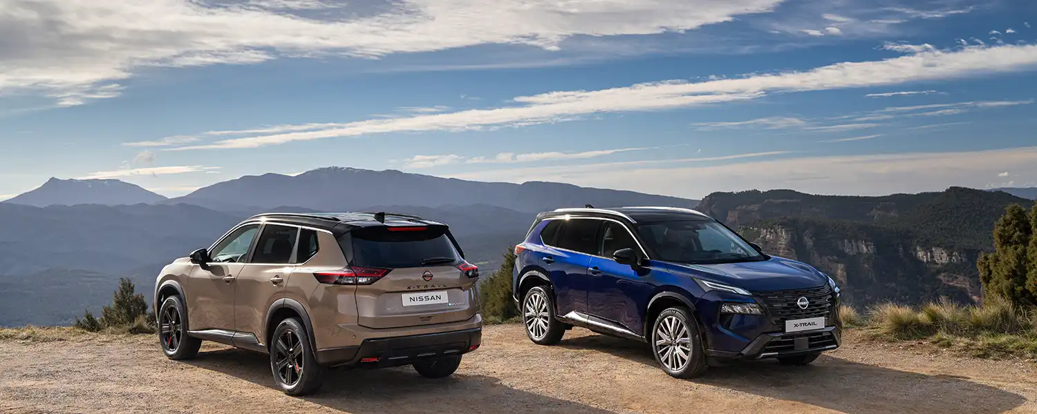 Two New 2026 Nissan X‑Trail SUVs parked on a dirt road with mountainous landscape and cloudy sky in background. Vehicles feature modern designs with N-Trek in Coastal Dune exterior color and Tekna in Sukomo Blue exterior body color, highlighting rugged outdoor capability and style.