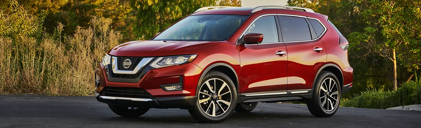 3/4 view of a red Nissan Rogue parked in front of the edge of a forest
