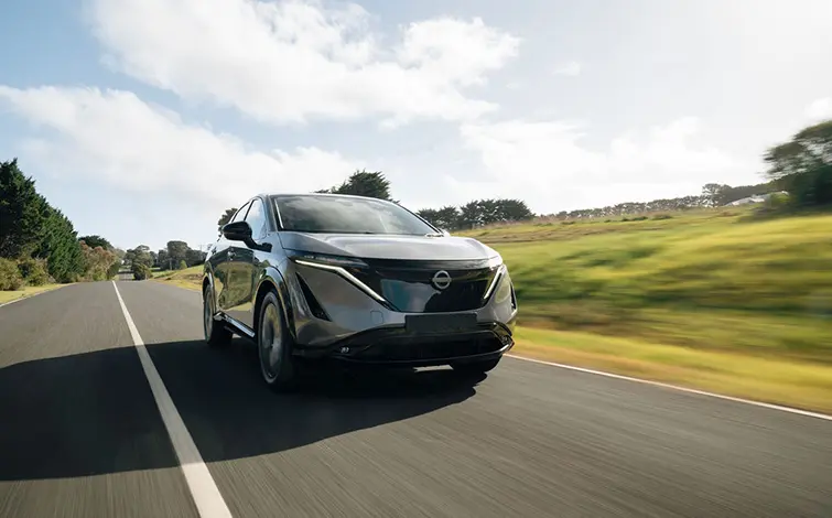 グレーの日産アリアが舗装された道路を前方右斜め向きに走行している。車両正面が低い位置から写され、背景には緑の草地と並木、青空に白い雲が広がっている。道路の白線が進行方向に伸びている。