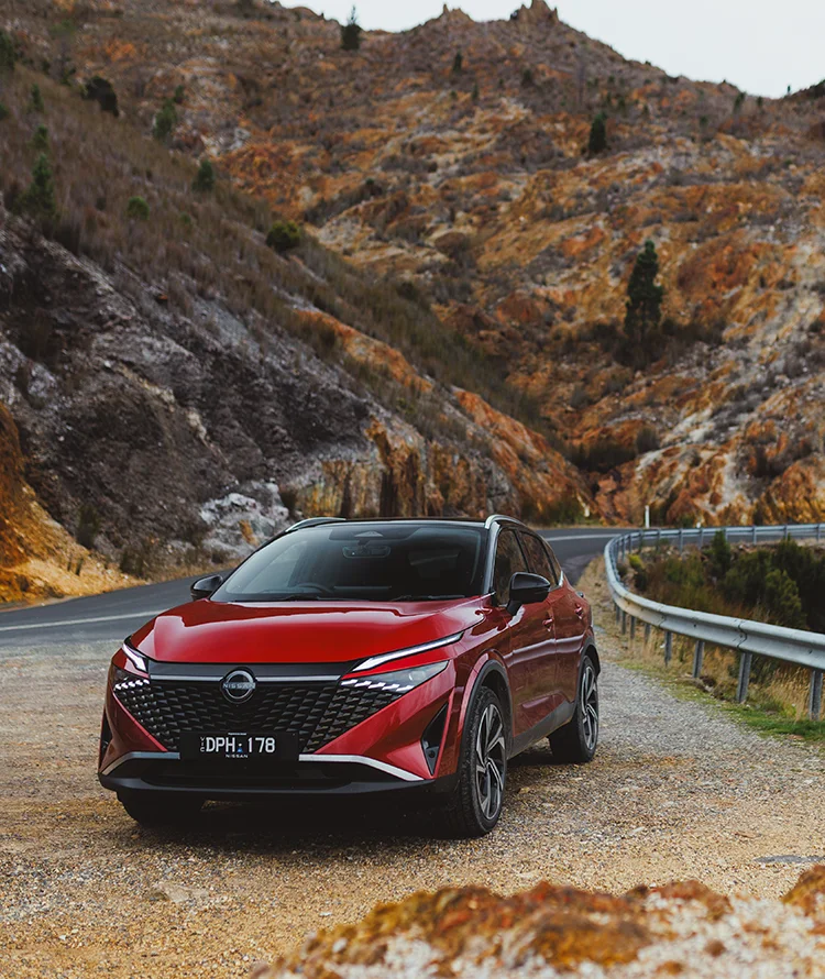 2026 Nissan Qashqai Ti-L E-POWER finished in Fuji Red with Black Roof parked beside a winding mountain road with rugged rock formations in the background.