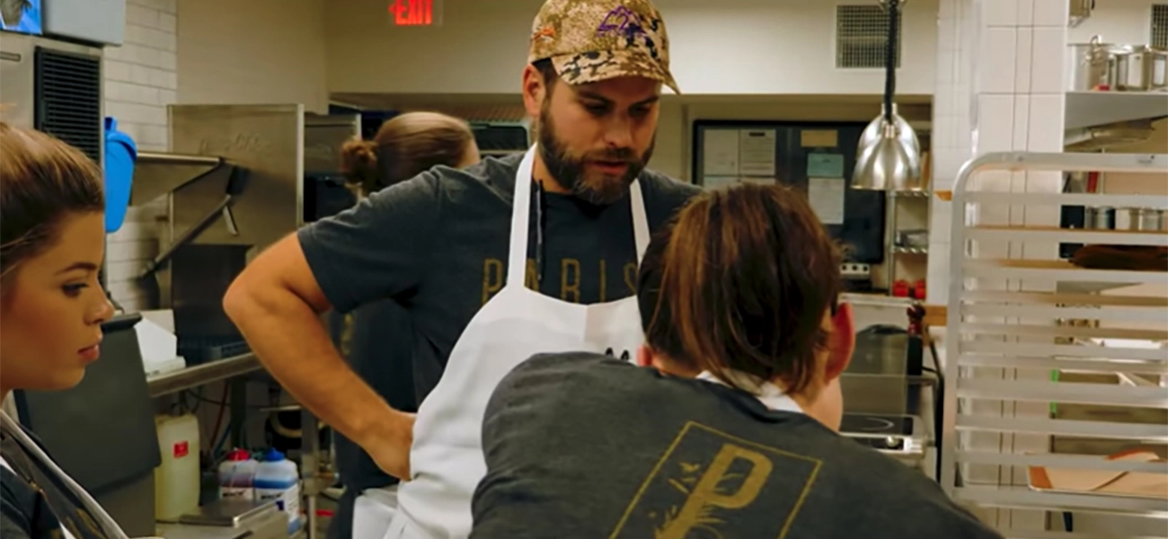 A man in white apron talking to his staff in a restaurant kitchen.