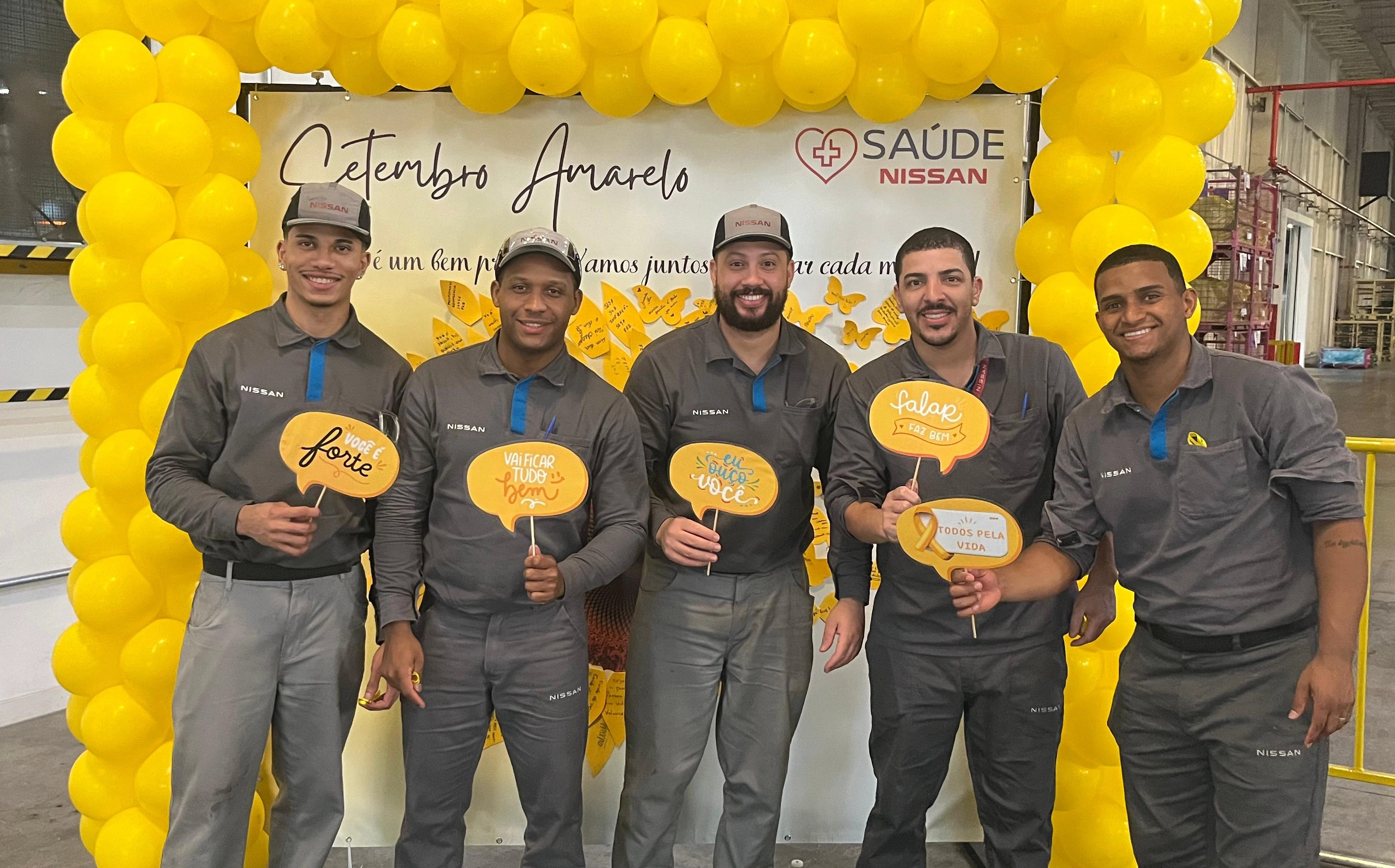 Quatro funcionários homens com uniforme da Nissan pousam para foto em frente a um painel do Setembro Amarelo com bolas amarelas no entorno.