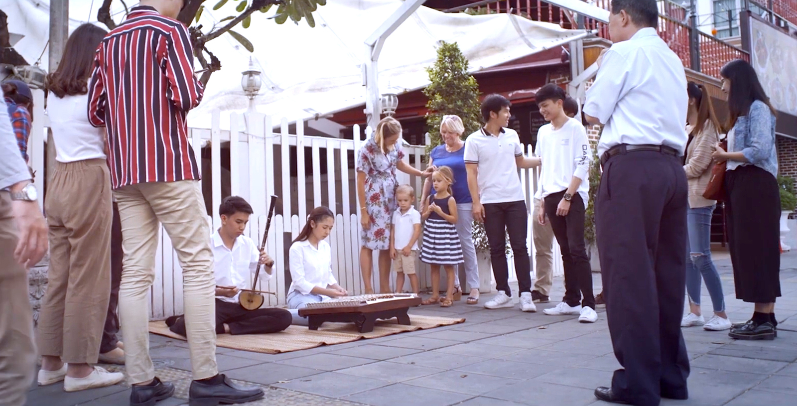 2 people playing musical instruments sitting on the ground while a group of people surrounding them.