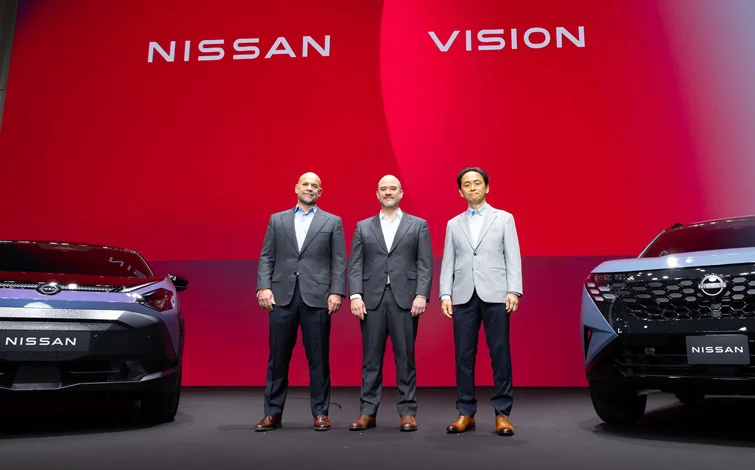 On a stage with a red background, three people are standing side by side facing forward, with Nissan vehicles positioned on the left and right, both facing front. The words “NISSAN” and “VISION” appear in the background.
