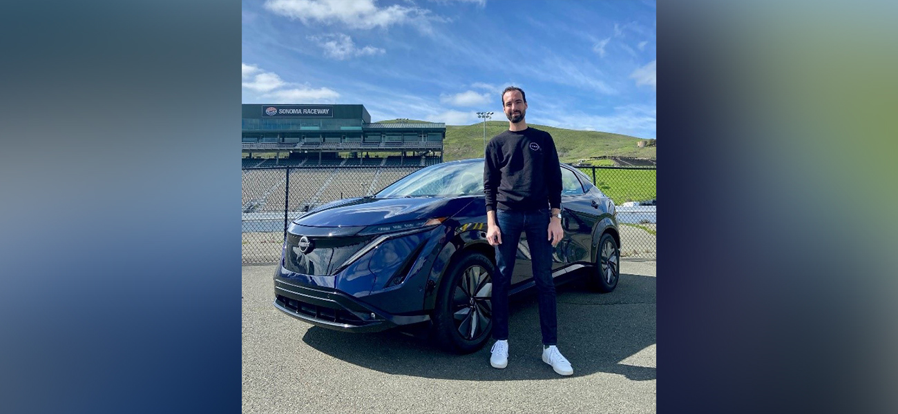 Man standing facing forward and smiling in front of blue vehicle near race track