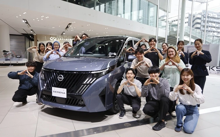 明るい自動車ショールーム内で、グレーのミニバン「日産セレナ」がやや左向きに展示され、その周囲に複数の人物が集まってポーズを取っている。背景にはガラス張りの展示空間や車両展示エリアが見える。