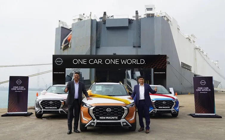 スーツを着た男性2人が新型日産マグナイトの前に立っています。背景にはインドから車を輸送する大型コンテナ船があり、他の車も2台見えます。