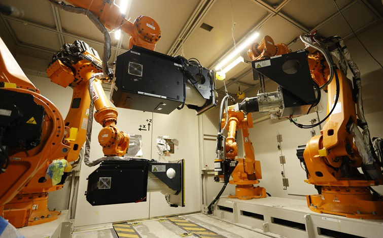 室内の作業エリアで、複数のオレンジ色の産業用ロボットアームが黒い装置を挟むように配置され、中央へ向けて稼働位置を取っている。背景は照明付きの天井と白い壁で、床には黄色と黒の注意ラインが引かれている。
