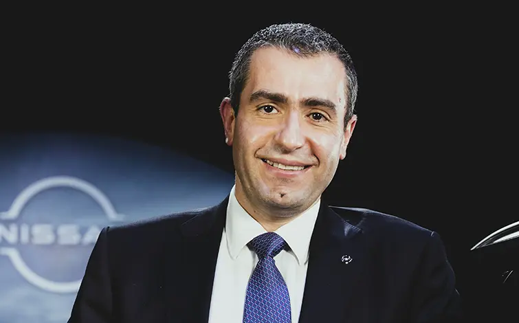 Photo portrait de Charbel Abi Ghanem, appuyé sur une voiture, costume foncé sur fond foncé avec logo Nissan flou derrière lui.