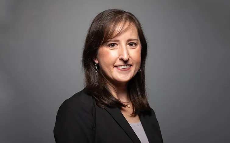 A woman with brown hair, smiling in a black suit against a gray background