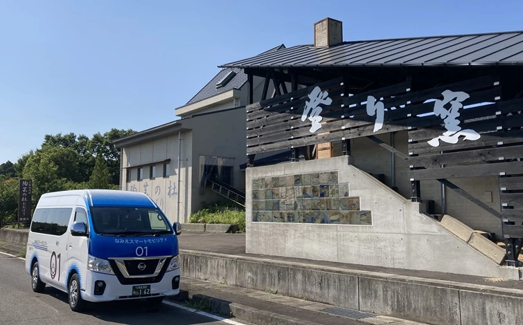 道路の左側を右向きに走行する白と青の「なみえスマートモビリティ」車両。右側には黒とグレーを基調とした建物があり、階段と看板が見える。背景には緑の木々と青空が広がり、地域の移動サービスを象徴する風景が撮影されている。