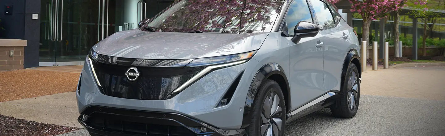 Front left 3/4 view of a gray 2024 Nissan Ariya parked under a tree with pink flowers
