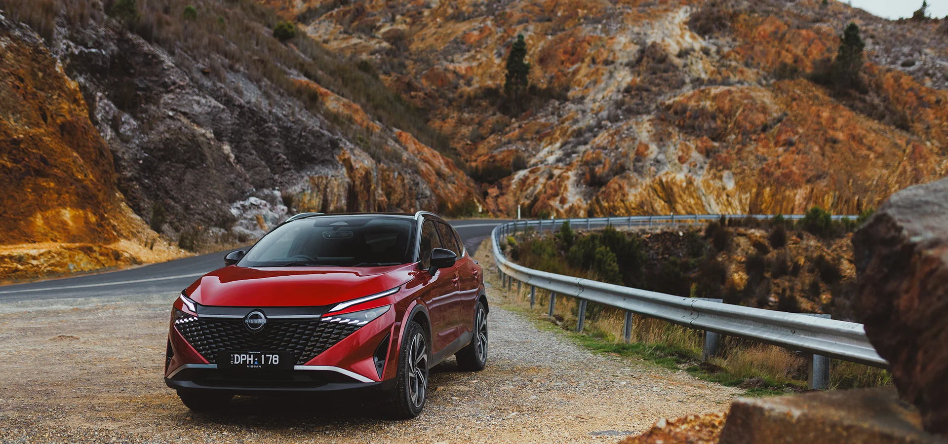 2026 Nissan Qashqai Ti-L E-POWER finished in Fuji Red with Black Roof parked beside a winding mountain road with rugged rock formations in the background.