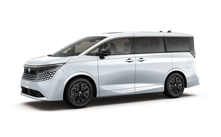 白いボディカラーの新型エルグランドを、左前方からの3/4アングルで撮影したスタジオ画像。車両は左向きに配置され、ブラックルーフとダークカラーのピラーが一体感のあるサイドビューを形成している。フロントには大型のパターングリルとシャープなヘッドランプを備え、ブラックのアルミホイールが足元を引き締めている。背景は淡いグレーの無地で、車両のみが際立つシンプルな構図。