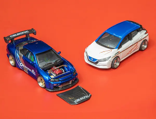 Duas miniaturas de carros sobre fundo vermelho: à esquerda, um esportivo azul com o capô removido exibindo o motor e a palavra "Calsonic"; à direita, um hatch branco e azul com adesivos nas laterais