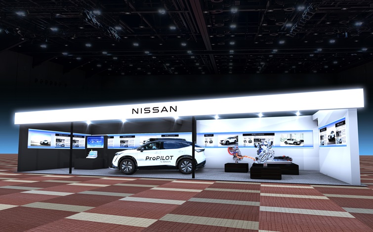 屋内展示会場に設置された日産の展示ブースの全景。正面上部に「NISSAN」のロゴがあり、中央には白い車両が正面左向きに展示されている。周囲にはモニターや展示パネルが並び、背景は暗い天井と照明、床は赤系のタイル模様になっている。