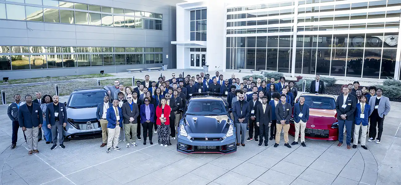 A large group of students, mentors and Nissan executives standing around a Nissan Ariya, GT-R and Z