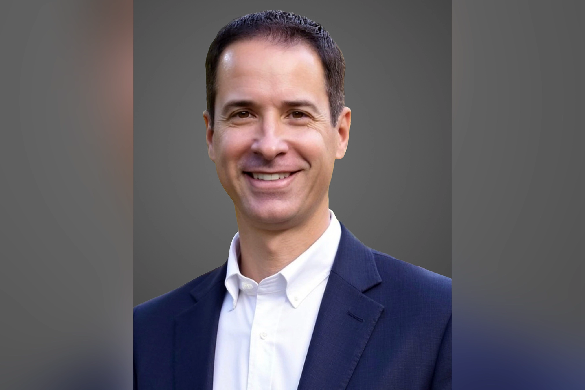 Andrew, smiling in a blue suit against a gray background.