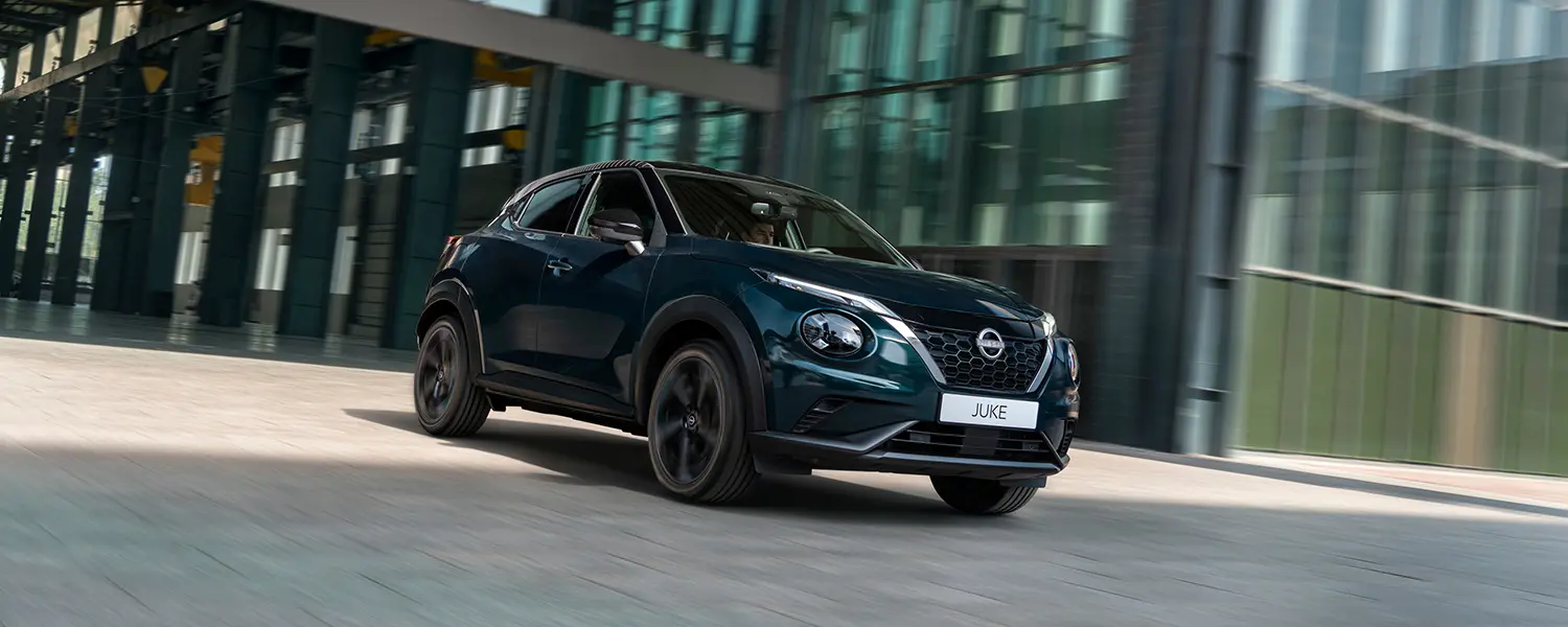 The Nissan JUKE Pulse driving through a modern urban setting, shown in motion with a sleek Ocean Deep exterior against glass-fronted buildings.