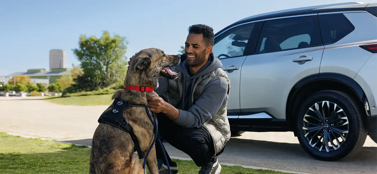 A man at a park, smiling and petting a large dog in front of a Nissan car.
