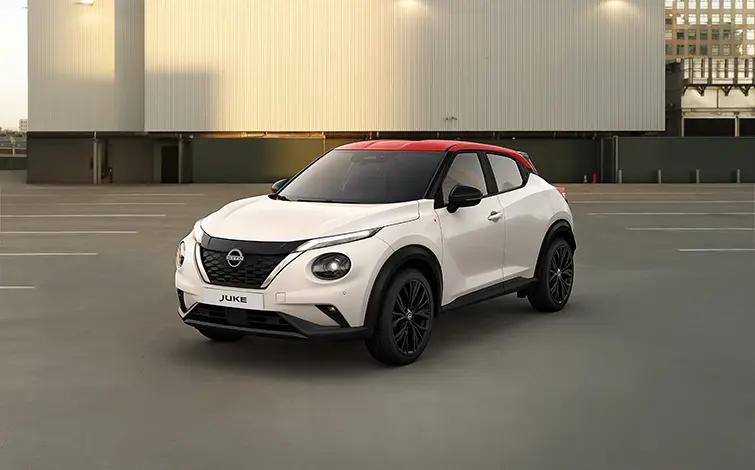 Blick von schräg vorne auf die Nissan Juke Redline Edition mit perlweißer Karosserie und Kontrastdach in Fuji Sunset Red, das Auto steht auf einem Parkplatz vor einem beleuchteten grauen Gebäude