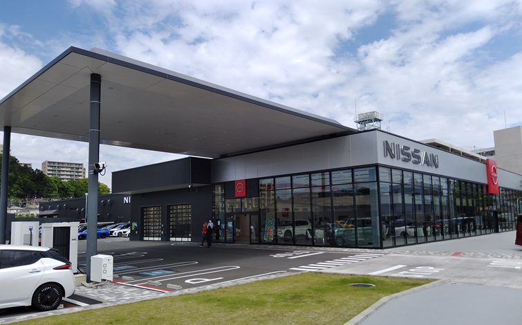 2025年10月より次世代型太陽光電池の実証実験を開始する日産神奈川販売株式会社 R1東戸塚店の店舗入口。青空の下、充電中の白いクルマや訪れる人たちがいる。