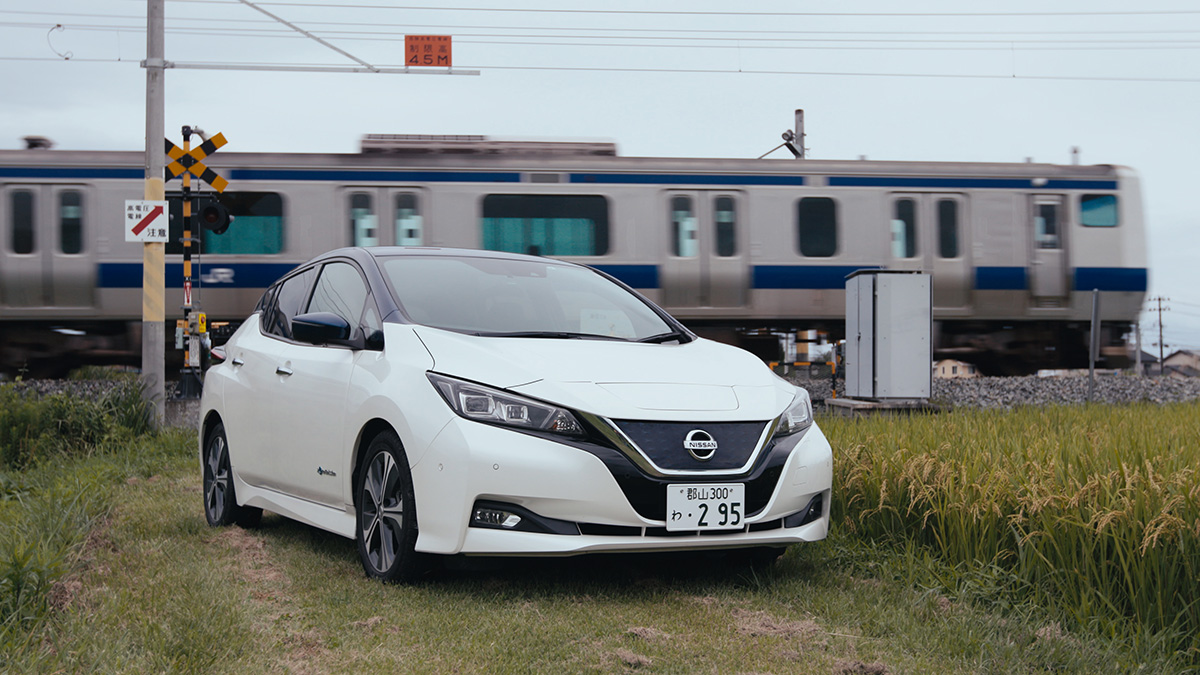 A white Nissan Leaf parked on a grassy road in front of a building