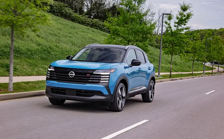 Front left 3/4 view of a blue 2025 Nissan Kicks driving along a road with vibrant grass and trees alongside.