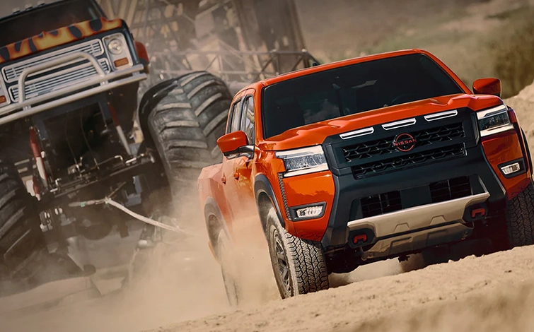 A 2026 Frontier driving along a dirt path, a monster truck follows.