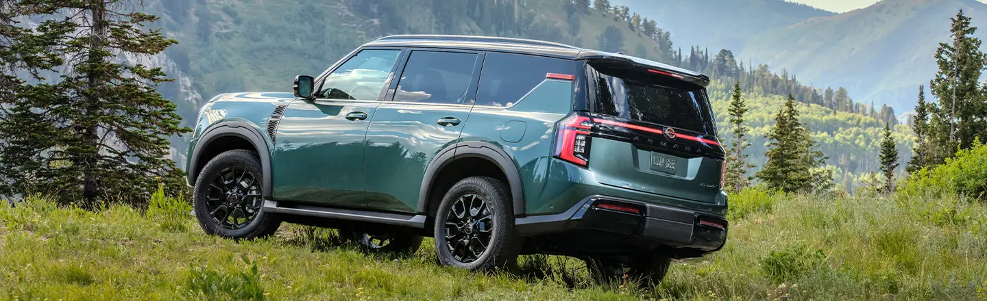 Left rear 3/4 view of a 2025 Nissan Armada parked on a grassy hilltop overlooking pine tree scattered mountains.