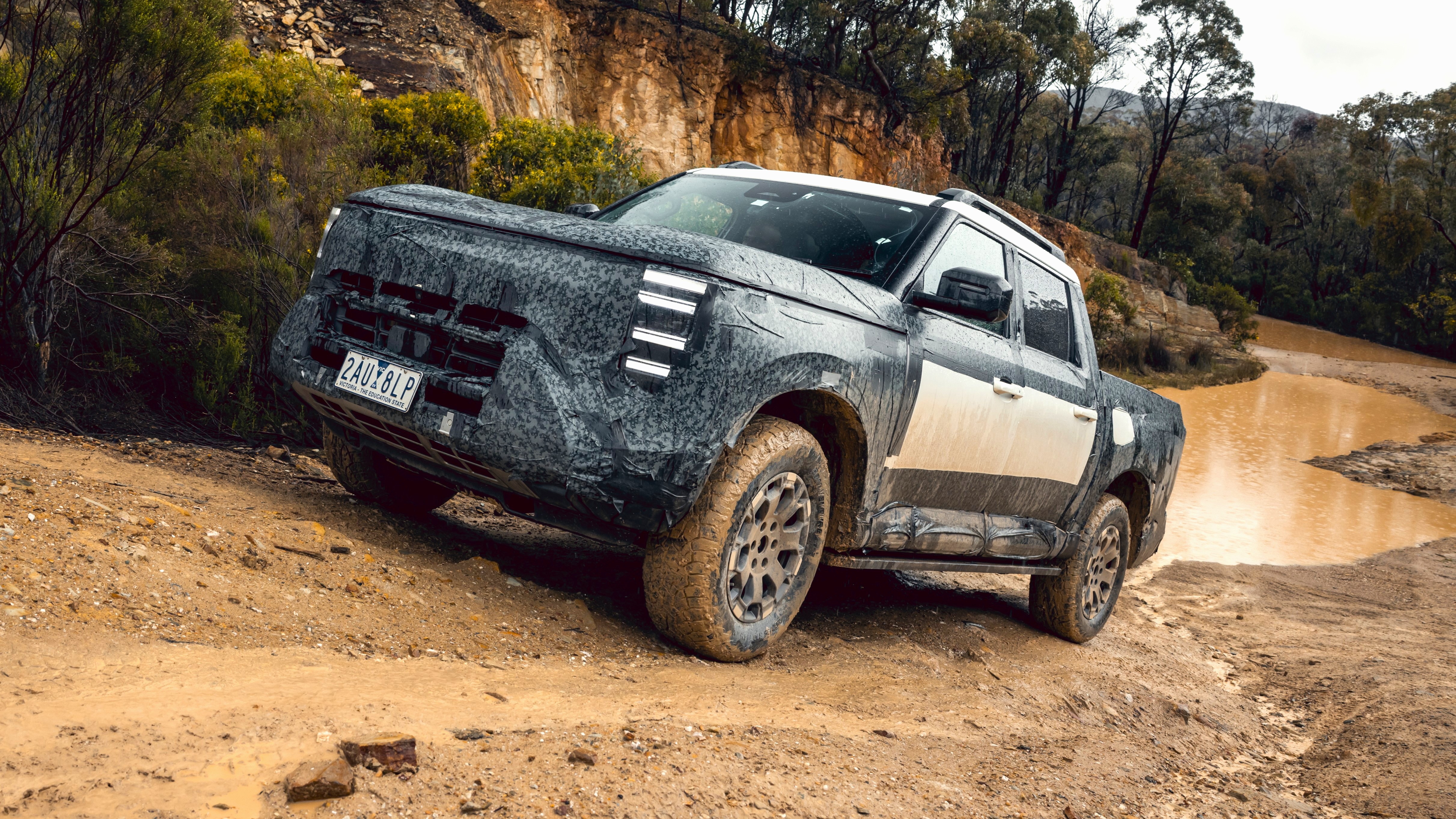 Front three-quarter of a 2027 Nissan Navara Pro PHEV dual-cab development vehicle traversing a muddy quarry track beside a water-filled crossing.