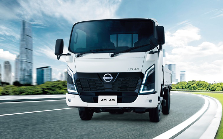都市部の道路を走行する白いアトラス。車両はカメラに向かって正面方向からやや斜めに進んでおり、フロントフェイスが強調されている。背景にはビル群と青空が広がる屋外の走行シーン。