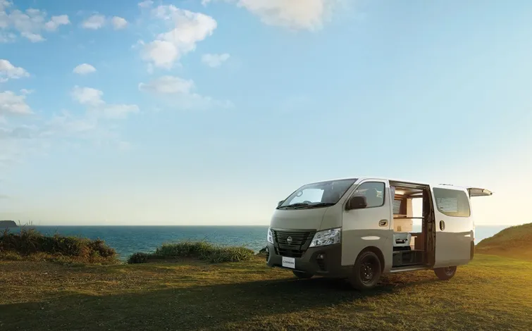 海沿いの草地に停車したキャニオンベージュのキャラバンMYROOM。右側面とフロントが見える角度で、リアゲートを開けている。背景には青空と海の水平線が広がり、柔らかな日差しが車体に当たっている。