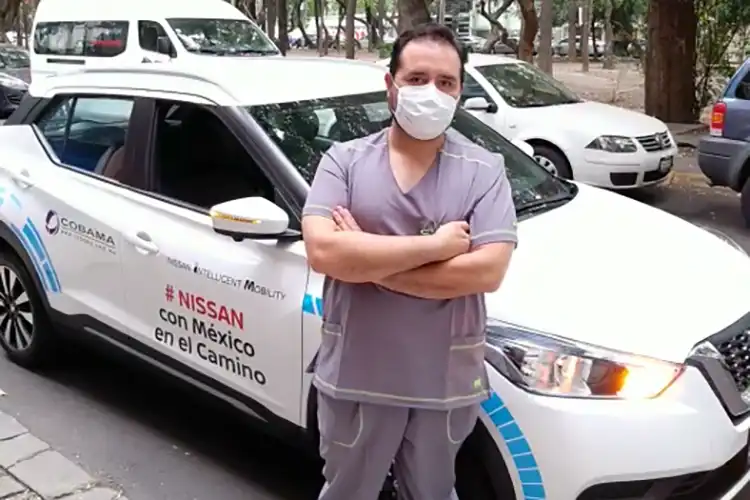 hombre con mascarilla bucal con brazos cruzados junto a un auto blanco
