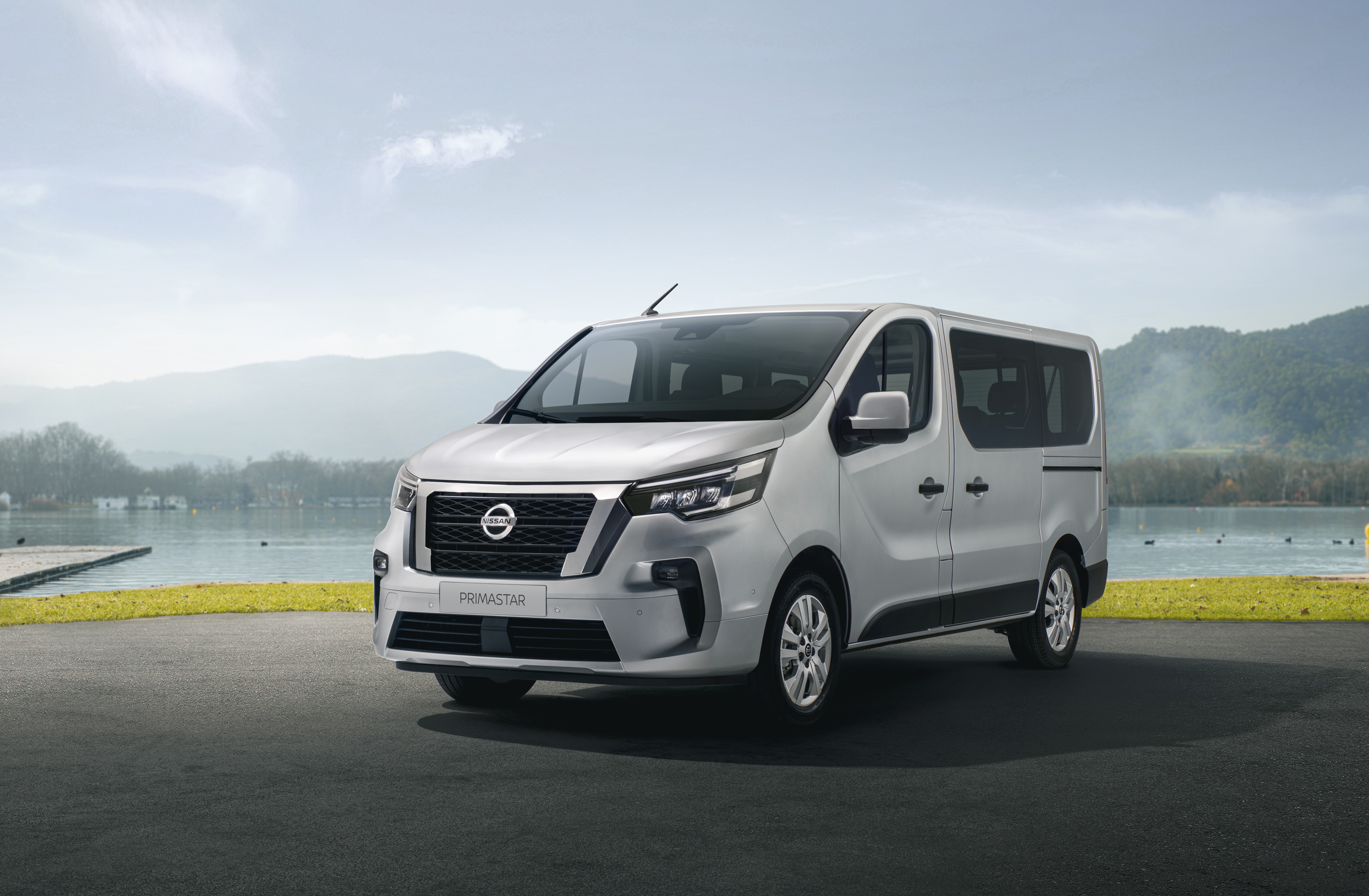 3/4 Frontansicht eines hellgrauen Nissan Primastar Kombi, der auf dunklem Asphalt vor einer Wiese und einem See steht