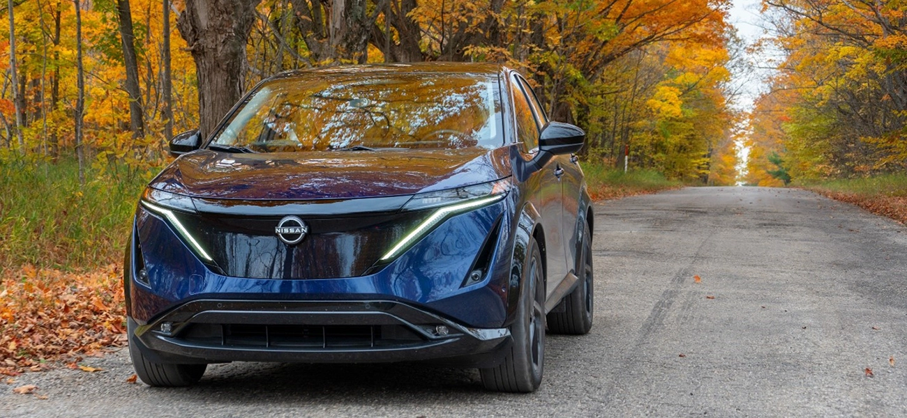A dark blue Nissan Ariya parked on a dirt road with bright orange turned tress lining the road behind.