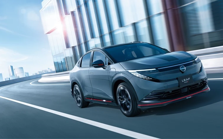 Front three-quarter view of a Leaf NISMO in NISMO Stealth Gray driving toward the left on an urban roadway. Glass-covered high-rise buildings form the background, while red NISMO accents highlight the vehicle's athletic styling.