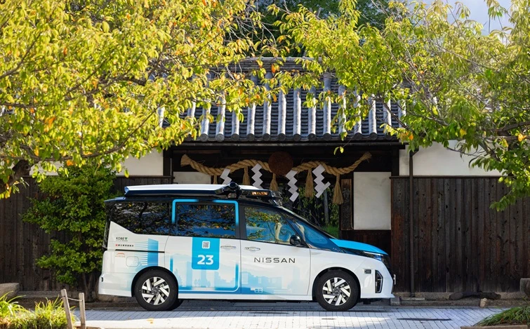 緑の木々に囲まれた和風建築の前に、左向きに駐車した白と青の車両が停まっている。晴天の屋外で、背景には木造の建物と植栽が見える。