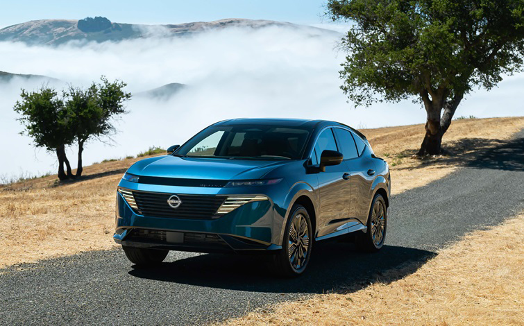 A blue Nissan Murano is shown from a front angled view driving on a road through a dry hillside landscape. Low clouds, distant hills, and a few solitary trees appear in the background.