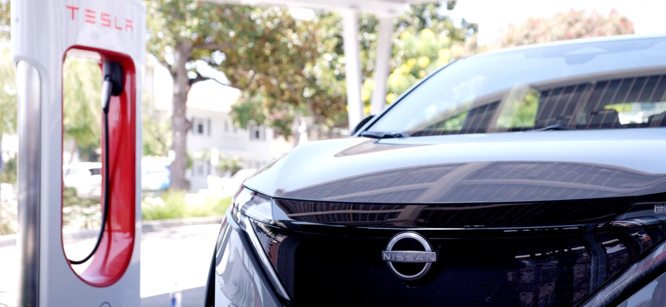 A front view of a gray 2025 Nissan Ariya parked by a charging station on a sunny day.