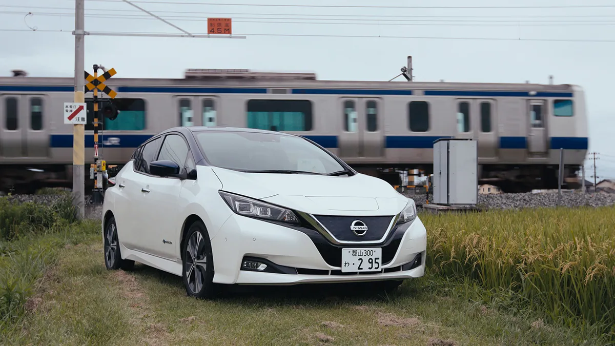 A white Nissan Leaf parked on a grassy road in front of a building