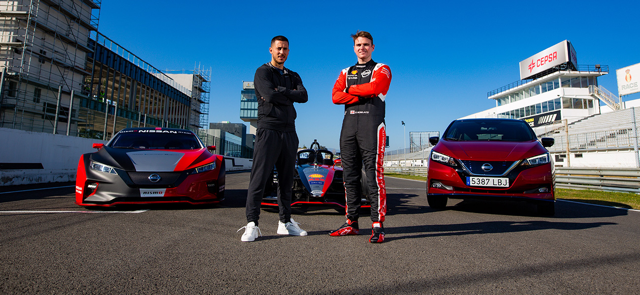 Two man standing with arms crossed in front of two cars on race track