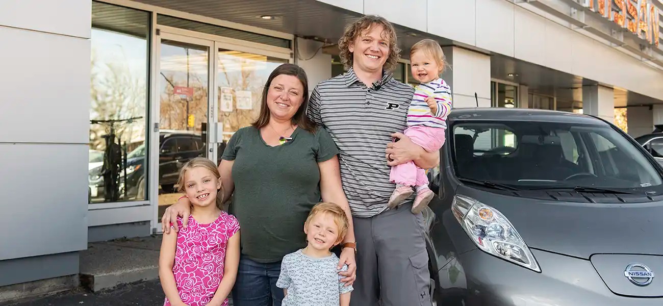 A family of 5 standing next to their gray Nissan LEAF.