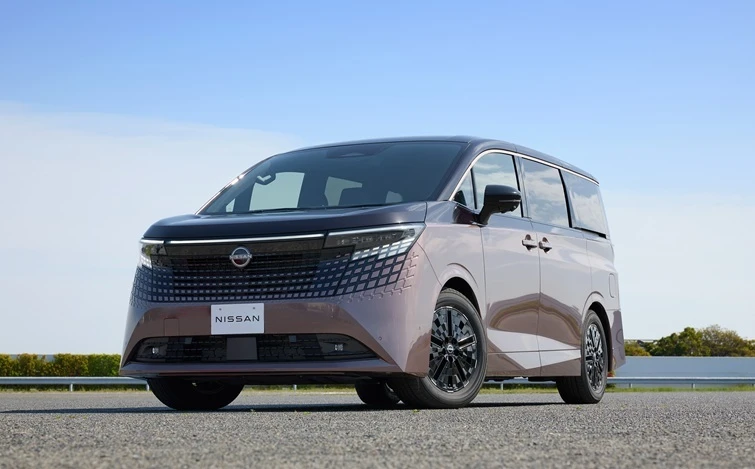青空を背景に、茶色を基調とした日産エルグランドが広々とした舗装路に斜め前向きで駐車している。