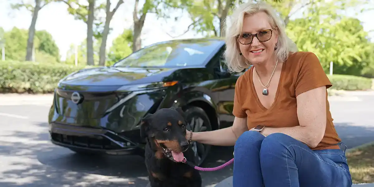 Nissan employee Henriëtte James sitting with her black and brown rescue dog, Tula, with a black Nissan Ariya and trees in the background