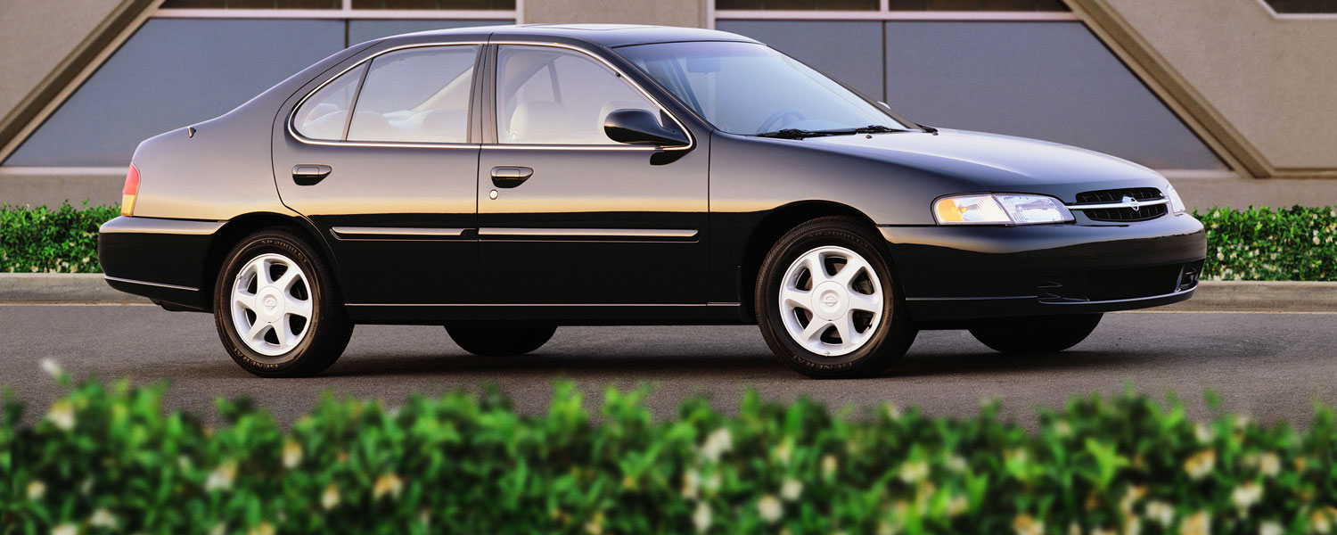 A black Nissan Altima parked on a paved road with small green bushes on the foreground.