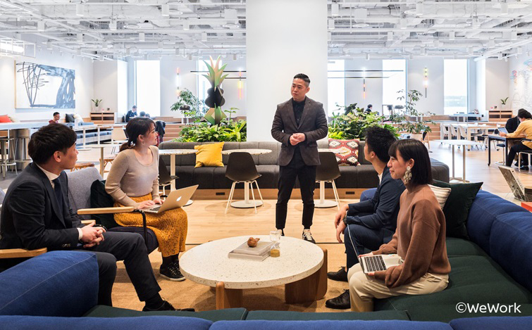 明るい色調のオープンオフィスで、観葉植物とソファに囲まれた円卓を中心に、前向きに座る数人がノートPCを使い、中央に立つ進行役を見ながら打ち合わせを行っている様子。