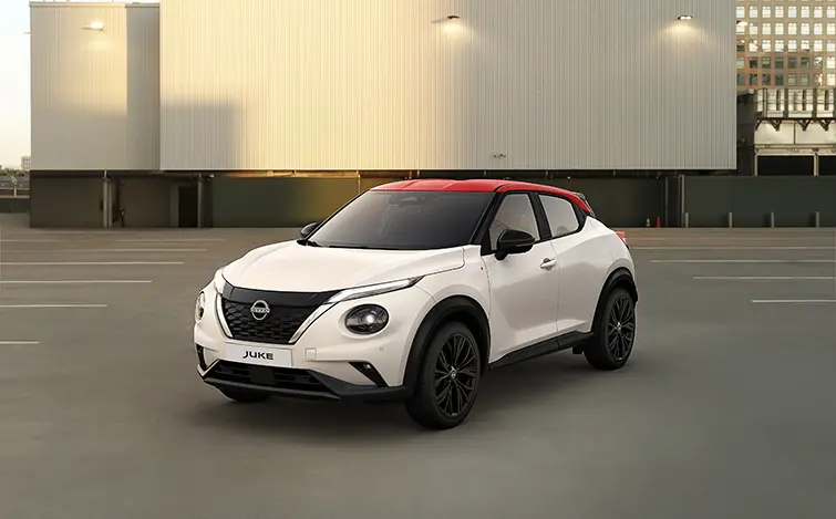 Blick von schräg vorne auf die Nissan Juke Redline Edition mit perlweißer Karosserie und Kontrastdach in Fuji Sunset Red, das Auto steht auf einem Parkplatz vor einem beleuchteten grauen Gebäude