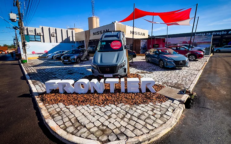 Imagem externa de um estande da Nissan com diversos carros expostos. Em destaque, uma picape Nissan Frontier cinza posicionada sobre uma área central com pedras, com letras tridimensionais brancas formando a palavra “FRONTIER”. Ao fundo, há mais veículos, o estande com o logotipo da Nissan e uma estrutura com lonas vermelha e branca que fornecem sombra parcial.